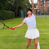 Woman wearing the Underscored Women's Ashdown Tank in White serving on a London grass tennis court, showing the racerback design and plastic-free performance fabric in movement