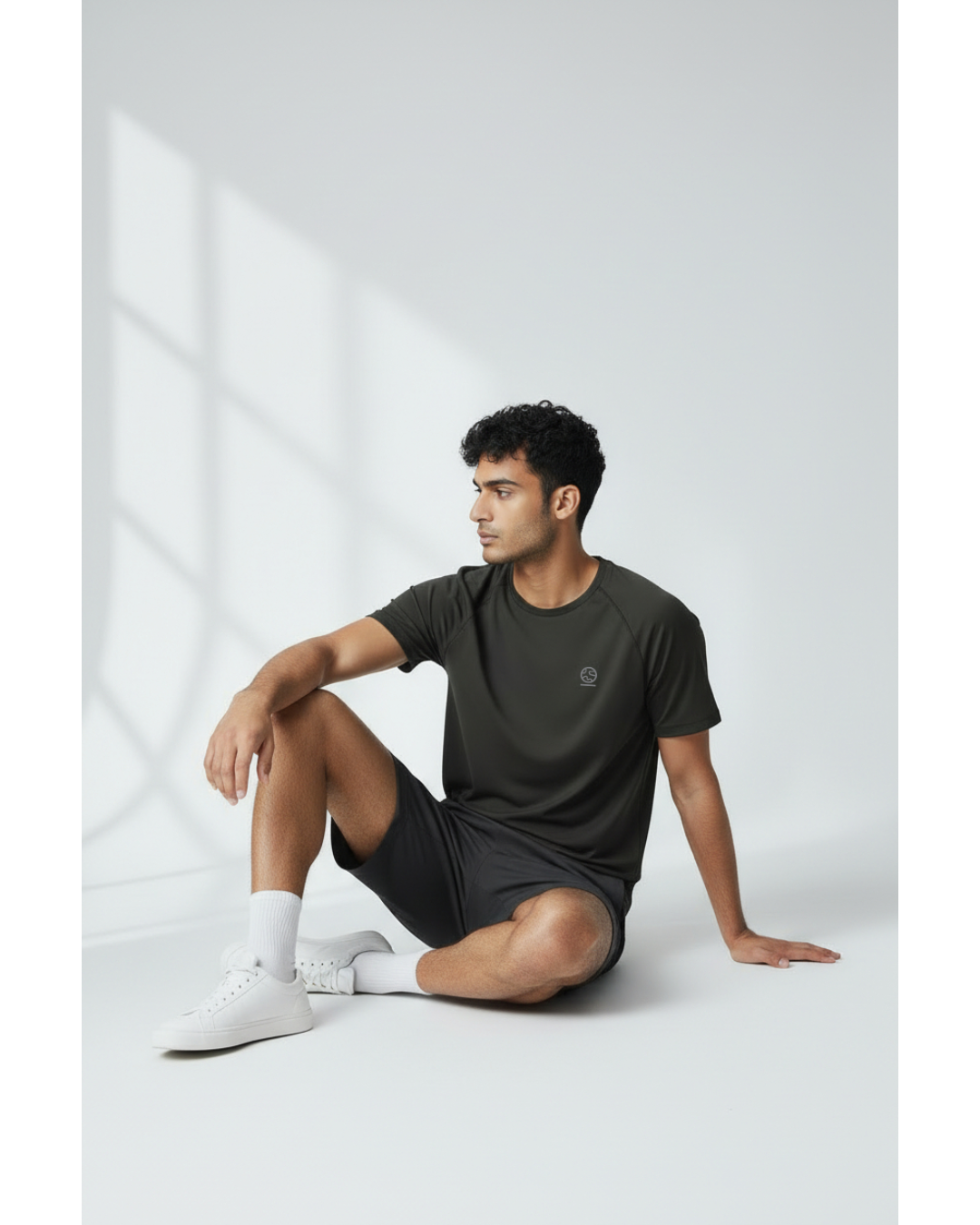 Man sitting on a white floor wearing a black underscored plastic-free t-shirt and grey shorts with a light grey background
