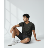 Man sitting on a white floor wearing a black underscored plastic-free t-shirt and grey shorts with a light grey background